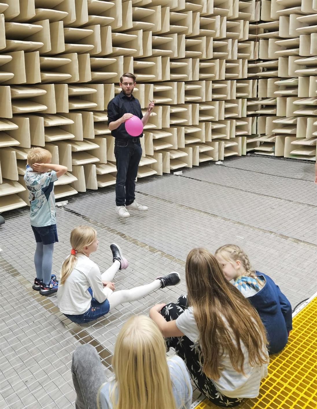 Ein Mitarbeiter des Instituts ISAVE der Hochschule Düsseldorf demonstriert Kindern bei „Türen auf mit der Maus“ ein Experiment mit einem pinken Luftballon in einem schallabsorbierenden Raum. Mehrere Kinder schauen zu, eines hält sich die Ohren zu.